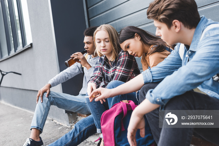 teenagers sitting, drinking beer from glass bottle and smoking cigarette