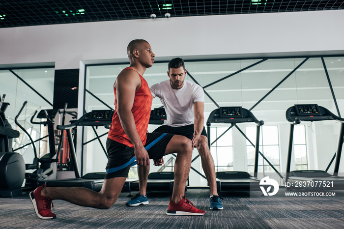 attentive trainer controlling african american sportsman exercising with resistance band