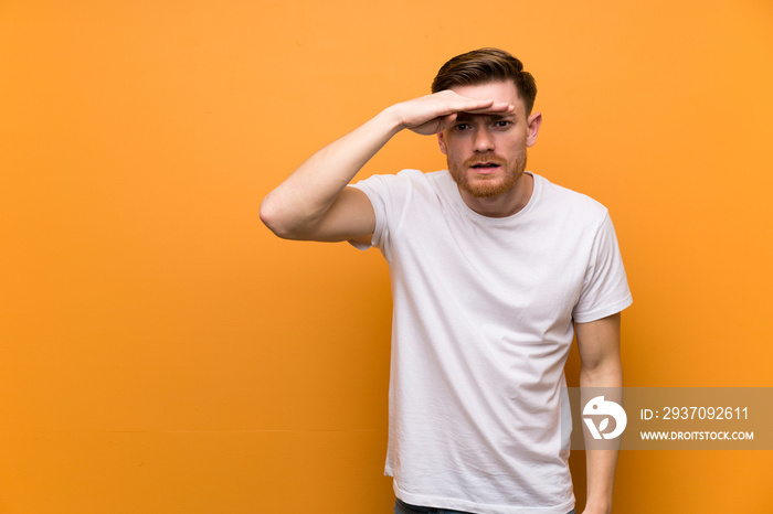 Redhead man over brown wall looking far away with hand to look something