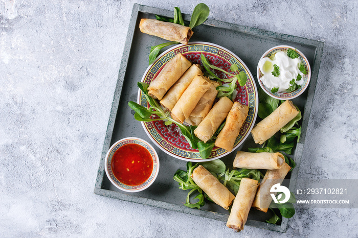 Fried spring rolls with red and white sauces, served in china plate on square wood tray with fresh g