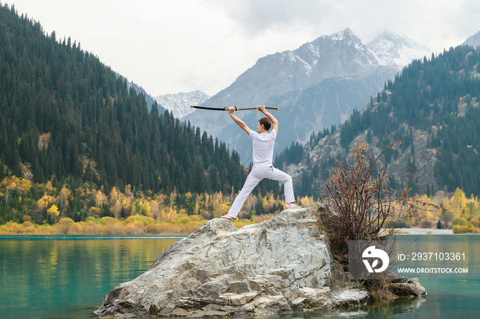 A man in white clothes with a sword stands on a large stone among the highlands.