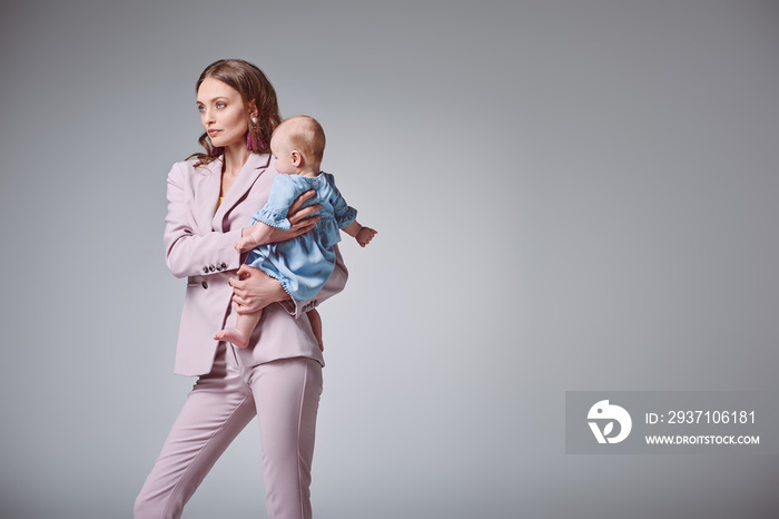 beautiful stylish young mother carrying adorable infant daughter and looking away isolated on grey