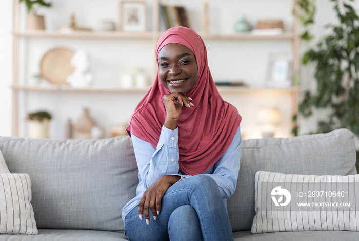 Joyful young african muslim woman in hijab posing on couch at home