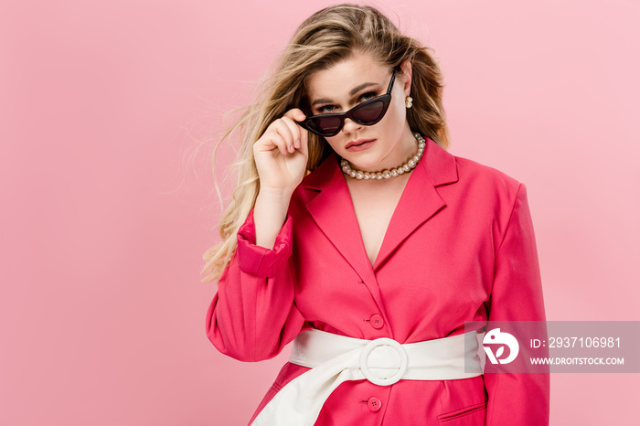 beautiful plus size woman in pink trench coat adjusting sunglasses and looking at camera isolated on