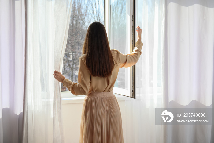 Pretty young woman opening window in room