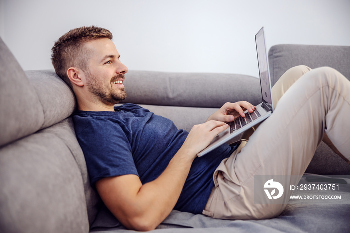 迷人的留着胡子微笑的男人躺在沙发上，用笔记本电脑上网的侧视图。
