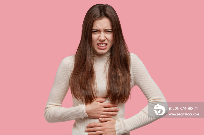 Photo of dissatisfied young woman keeps hands on belly, suffers from pain, has stomachache, displeas