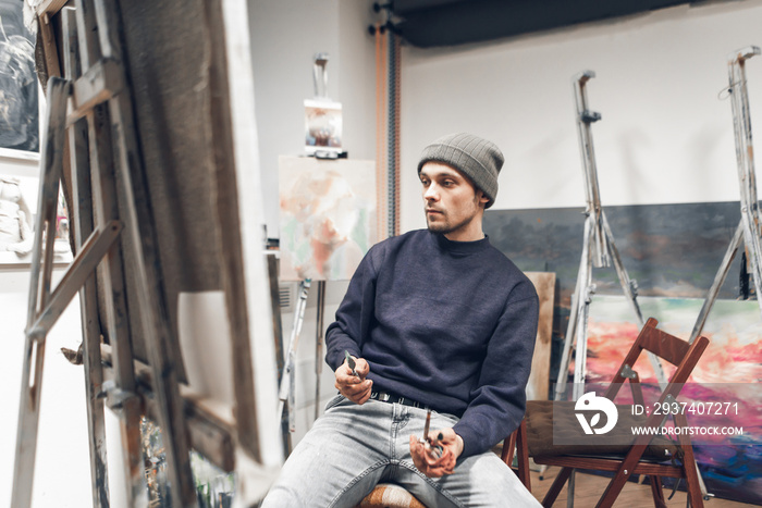 Young professional artist sits on a chair near an easel with a canvas, holds a brush in his hand and