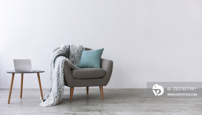 Modern armchair with blue pillow and soft plaid, next to small table with laptop in scandinavian int