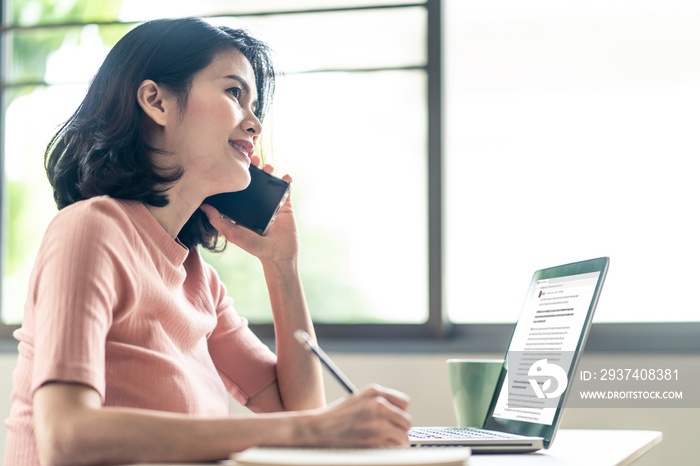 Asian young business woman sit and write note in a notebook while talk on phone and open files in la