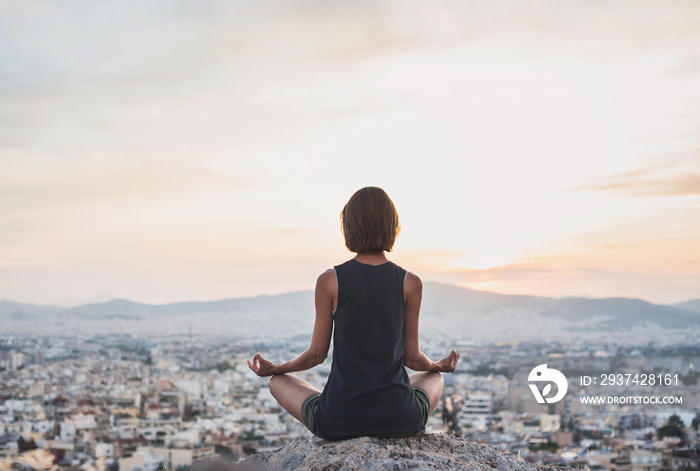 Young woman practicing yoga outdoors at sunset with a big city at the background. Harmony and medita