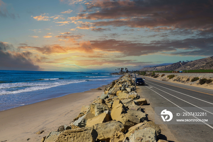 a shot of the beach near a long stretch of road with cars and large rocks along the beach, vast blue