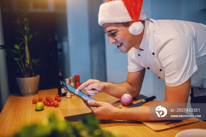 Handsome caucasian smiling chef in white uniform and with santa hat on head using tablet while leani