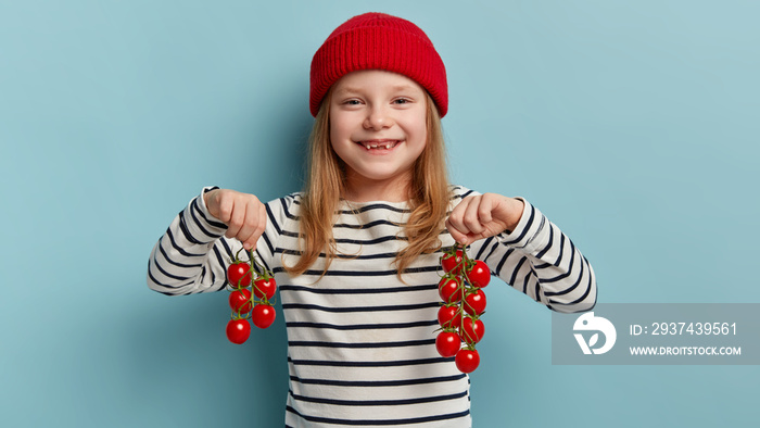 收获蔬菜的概念。可爱的小孩戴着红色帽子，穿着条纹毛衣，背着红色的雪