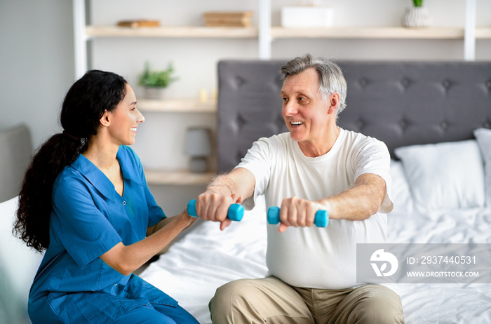 女性理疗师帮助积极的老人在退休时在床上用哑铃锻炼