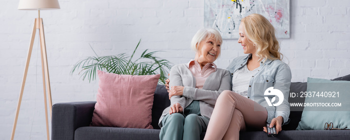 Senior woman smiling at daughter with remote controller on couch, banner