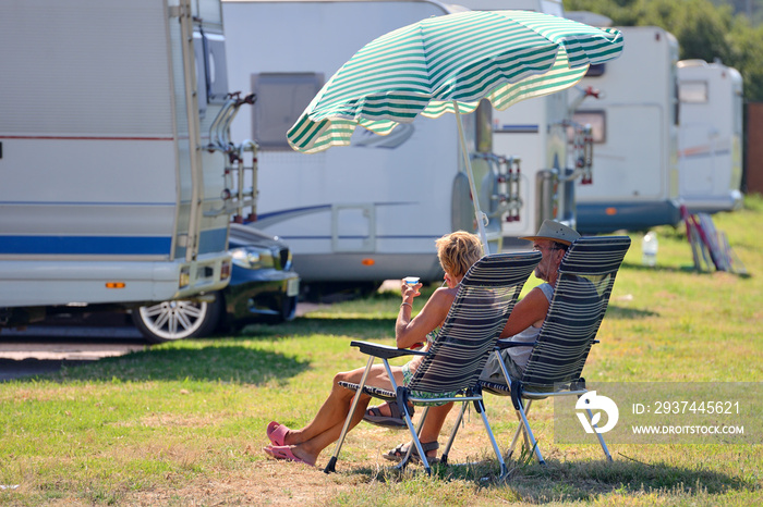 elderly couple is resting on chairs under a beach umbrella near a caravan