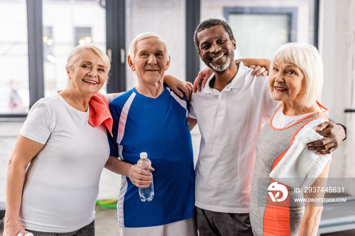 joyful multicultural senior sportspeople looking at camera and embracing each other at gym