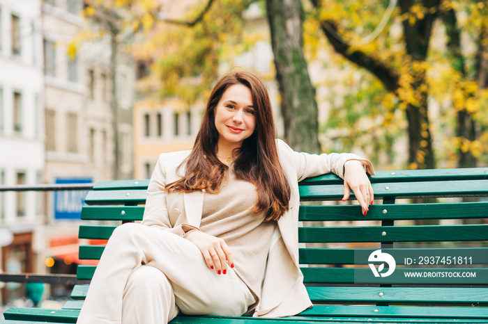 Outdoor portrait of beautiful woman resting in park, sitting on bench, wearing beige suit