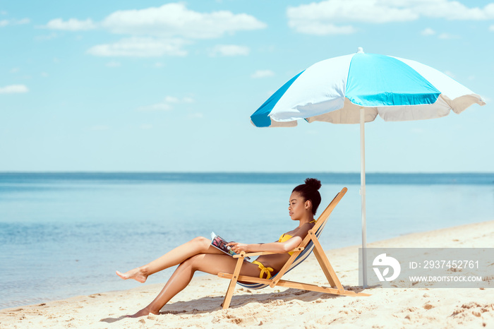 side view of african american woman reading magazine and relaxing on deck chair under beach umbrella