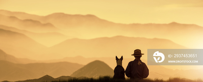 a silhouette Girl sits on top of a Mountain with a dog during a golden sunset.