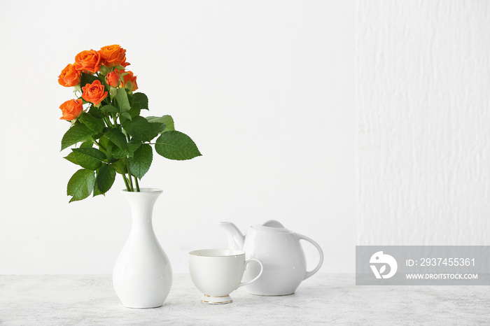 Vase with beautiful orange roses, teapot and cup on table against light wall