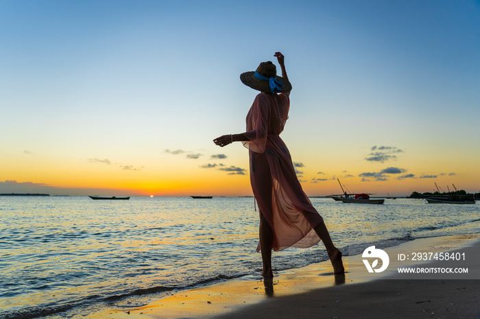 坦桑尼亚桑给巴尔岛日落时，海滩上戴着草帽和帕莱奥的美丽女孩