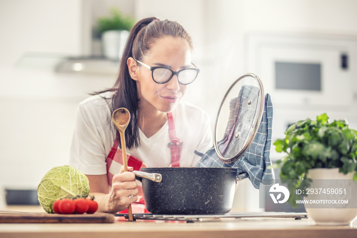 Woman holding spoon and lid is cooking at home, opening saucepan that fogs her glasses