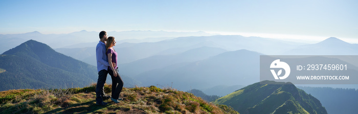 Full length of couple in love standing on grassy hill and looking at beautiful mountains. Young man 