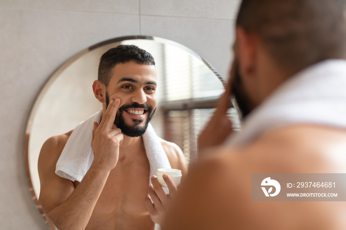 Handsome guy looking in the mirror applying cream on face