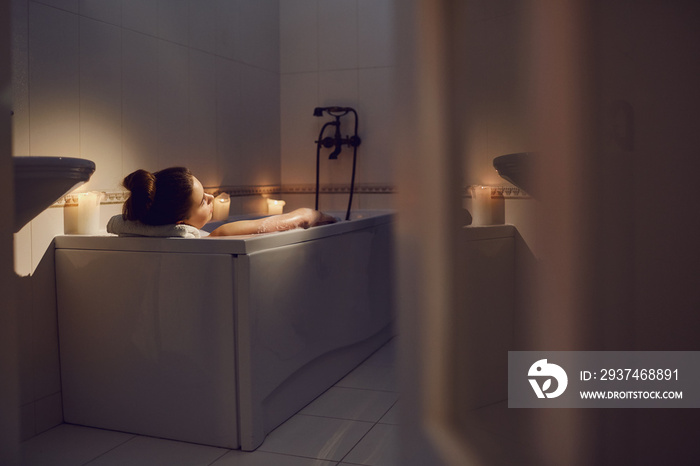 Young girl is resting in a bathtub with foam in bethroom with candles.