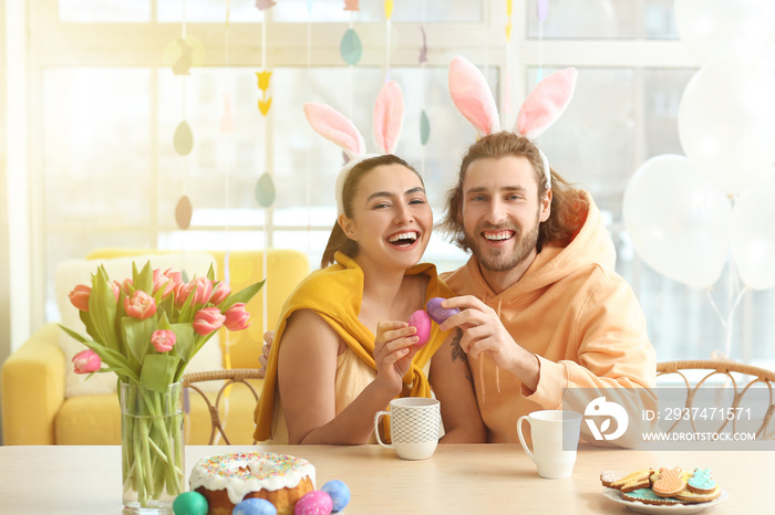 Happy couple in bunny ears cracking Easter eggs at table