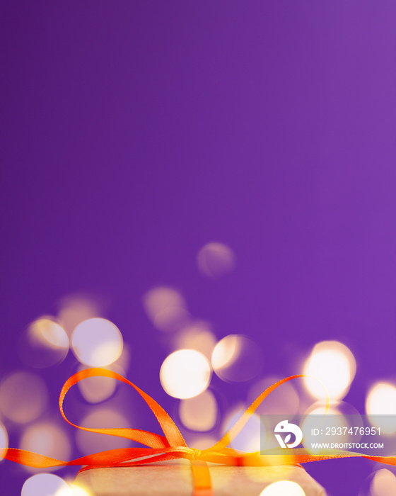 Close up of an orange satin ribbon bow at the bottom with golden bokeh on vibrant purple blurred bac