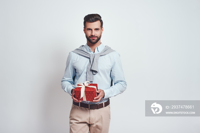 Surprise! Charming bearded man is holding a gift box and smiling while standing in studio on a grey 