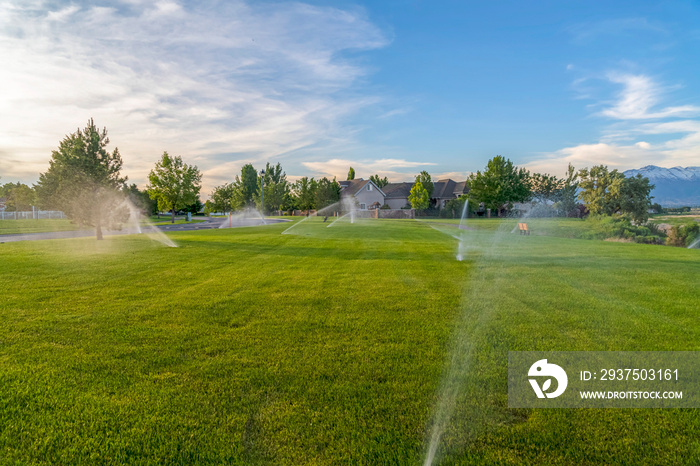 Sprinklers spraying water on green grasses with homes mountain and blue sky view