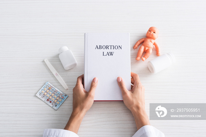 top view of woman holding book with abortion lettering near baby doll and abortion pills