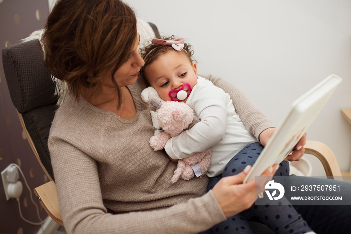 High angle view of mother reading book for cute daughter while sitting on chair at home