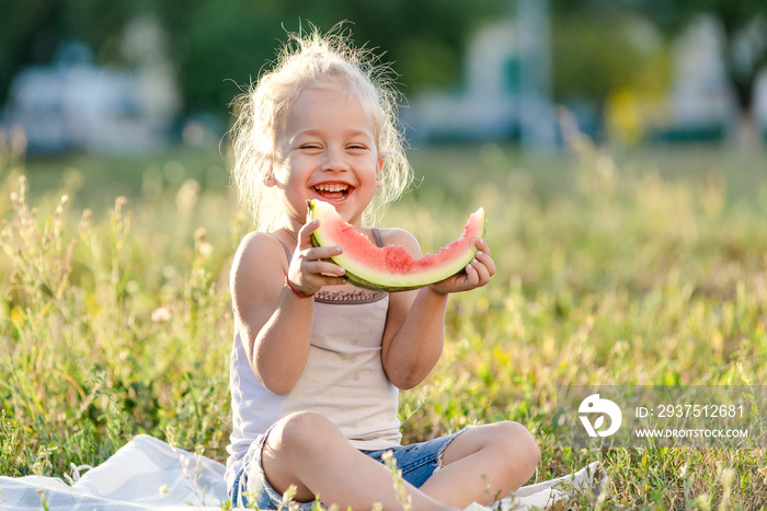 Little blond girl eating watermelon in the park.