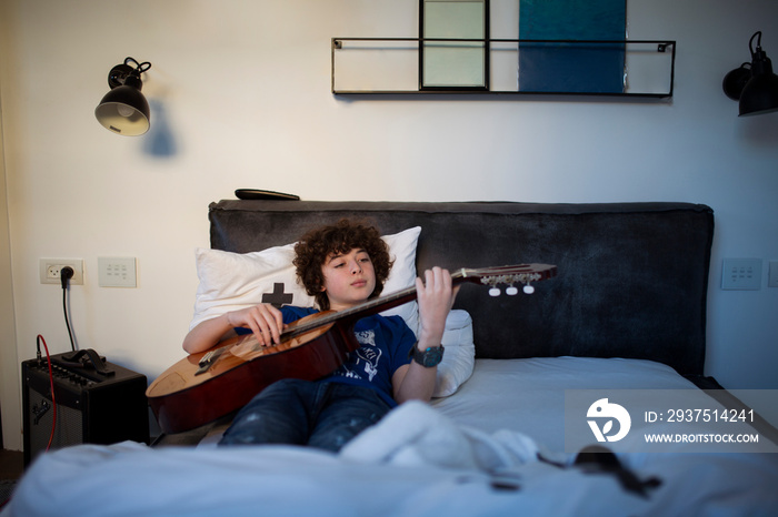 Cute boy playing guitar while relaxing on bed against wall at home