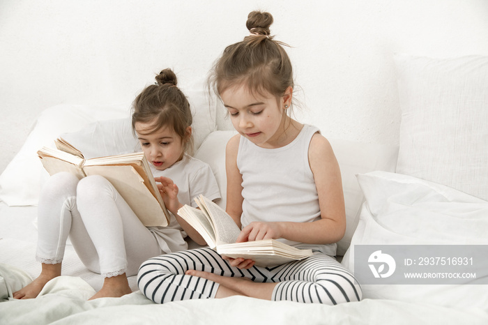 Two cute little sister girls are reading a book on the bed in the bedroom.