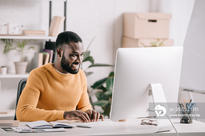 smiling handsome african american designer in orange sweater working at computer in office