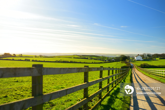 Fence casting shadows on a road leading to small house between scenic Cornish fields under blue sky,