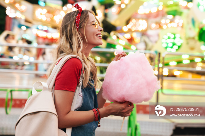 Image of blonde charming woman eating sweet cotton candy while walking in amusement park