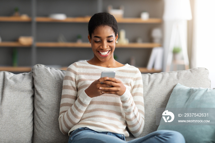 Cheerful Black Lady Relaxing With Smartphone In Living Room, Messaging With Friends