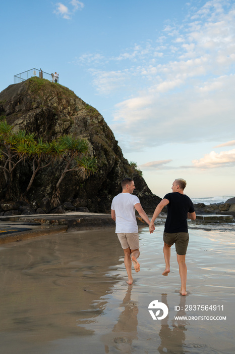 一对同性恋情侣手牵着手在有树和石头的海滩上奔跑