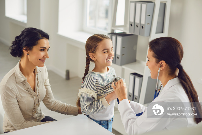 A woman pediatrician makes a setoscope examination to a child girl with mom in a clinic office.