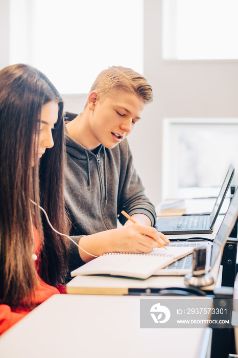 Male and female students writing in book while using laptop at desk