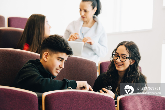 Teenage friends using mobile phone in class while professor standing in background