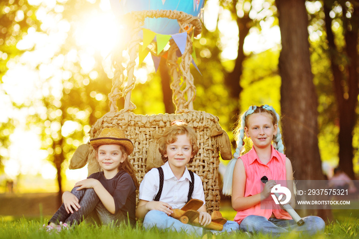 three small preschool children sit on the grass against the background of a basket of blue balloons 