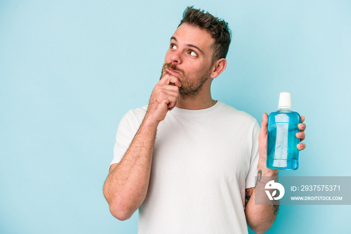 Young caucasian man holding mouthwash isolated on blue background looking sideways with doubtful and